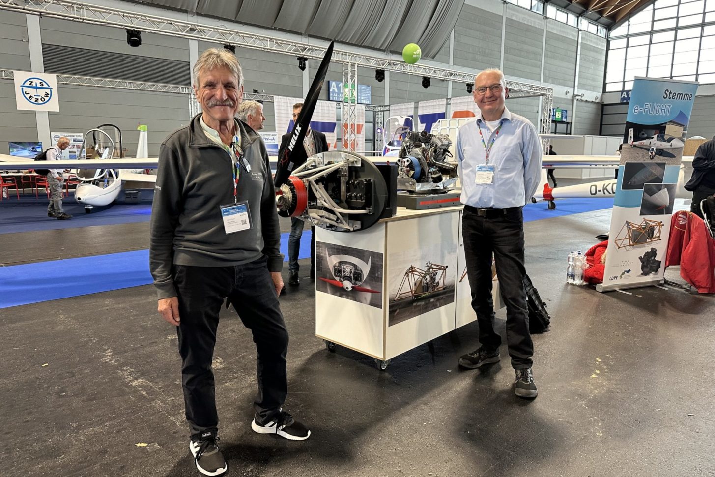 Klaus Ohlmann (links) und Karl Pickan (rechts) präsentieren vor einem Modell des innovativen Elektroantriebs, bestehend aus Batterien und einem Range Extender. Dieses Konzept verkörpert die Zukunft des emissionsarmen Segelfliegens, kombiniert traditionelle Glidertechnologie mit moderner elektrischer Effizienz.